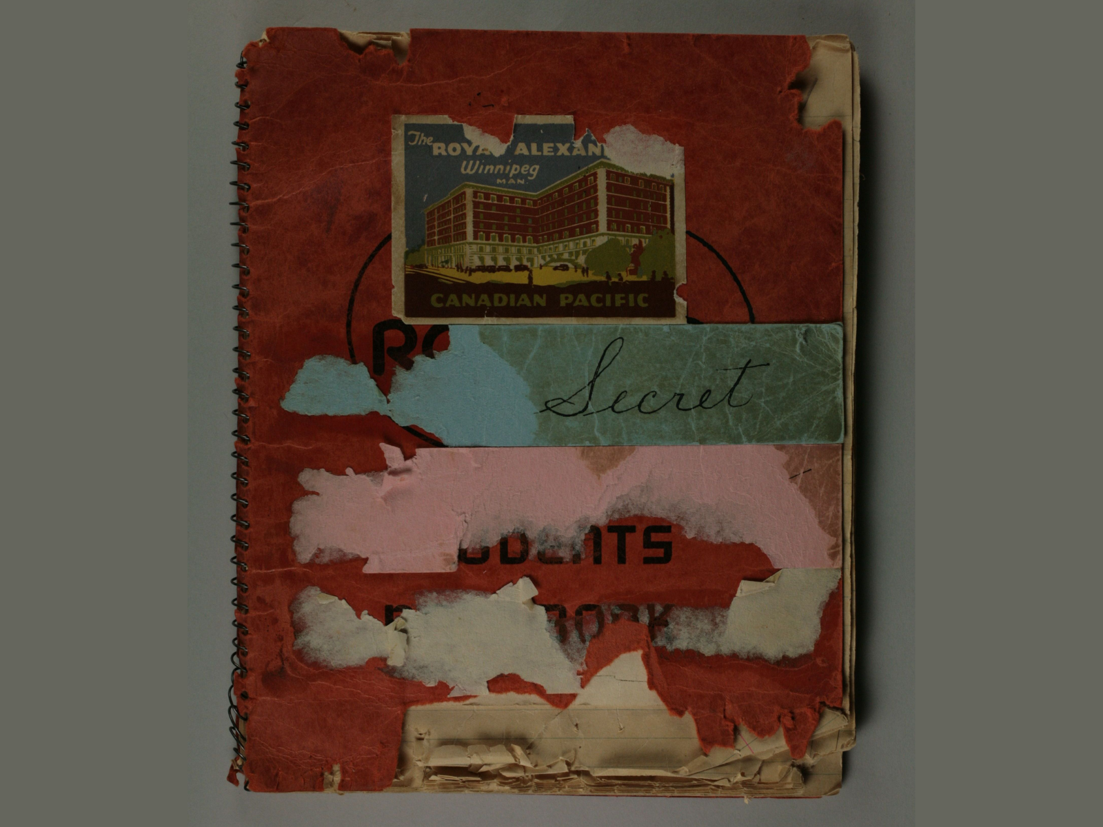 A worn sketchbook, it’s red cover torn and with the remains of torn off tape or stickers. A reminding piece of blue tape reads, “Secret” below a slightly torn sticker showing an image of a large red and white multi-story building and text reading, “The Royal Alexander / Winnipeg Man. / Canadian Pacific”.