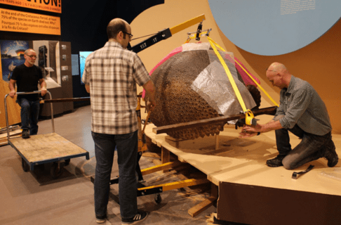 Three individuals work together to move a large Glyptodont carapace onto a wheeled cart from a platform using an engine hoist lift.