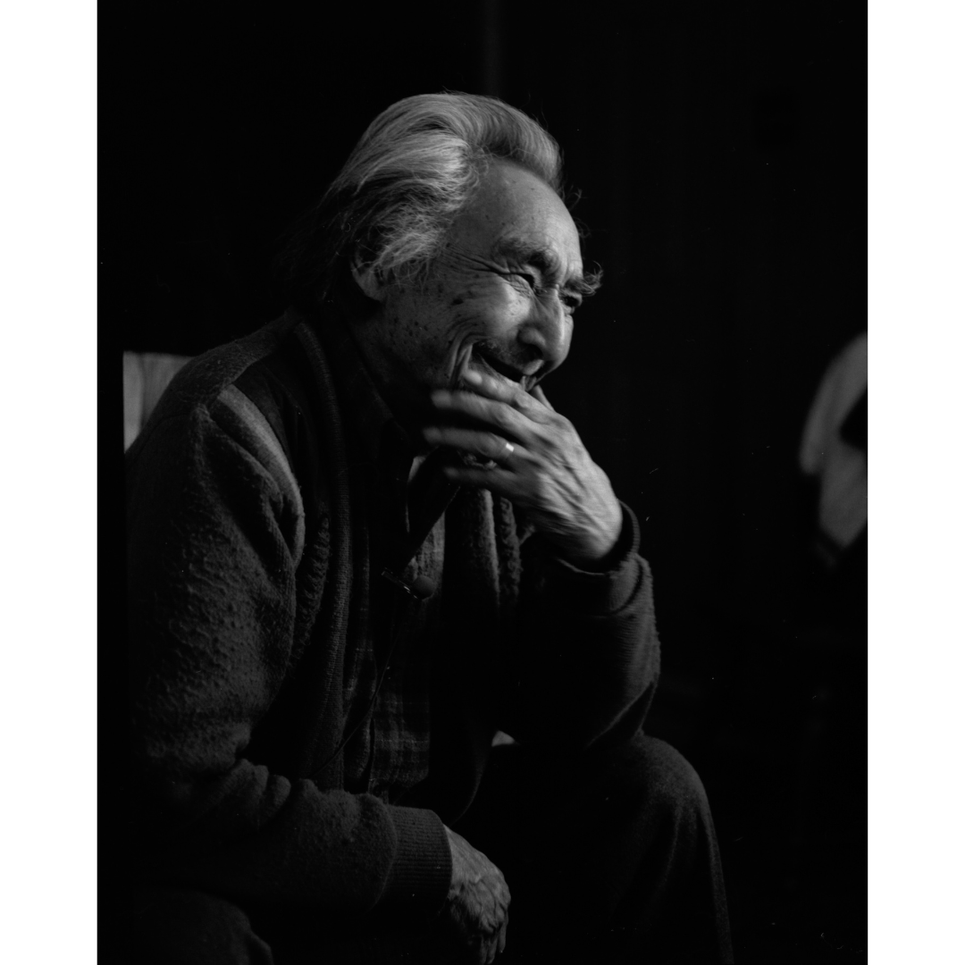 A black and white portrait of a seated older man from the side, smiling and laughing behind his hand.