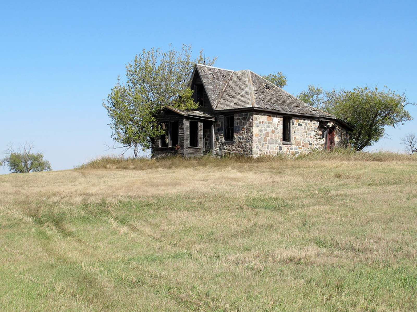 View up a gentle hill towards a now abandoned homestead built out of fieldstone.Overgrown bushes and trees grow close to the building.