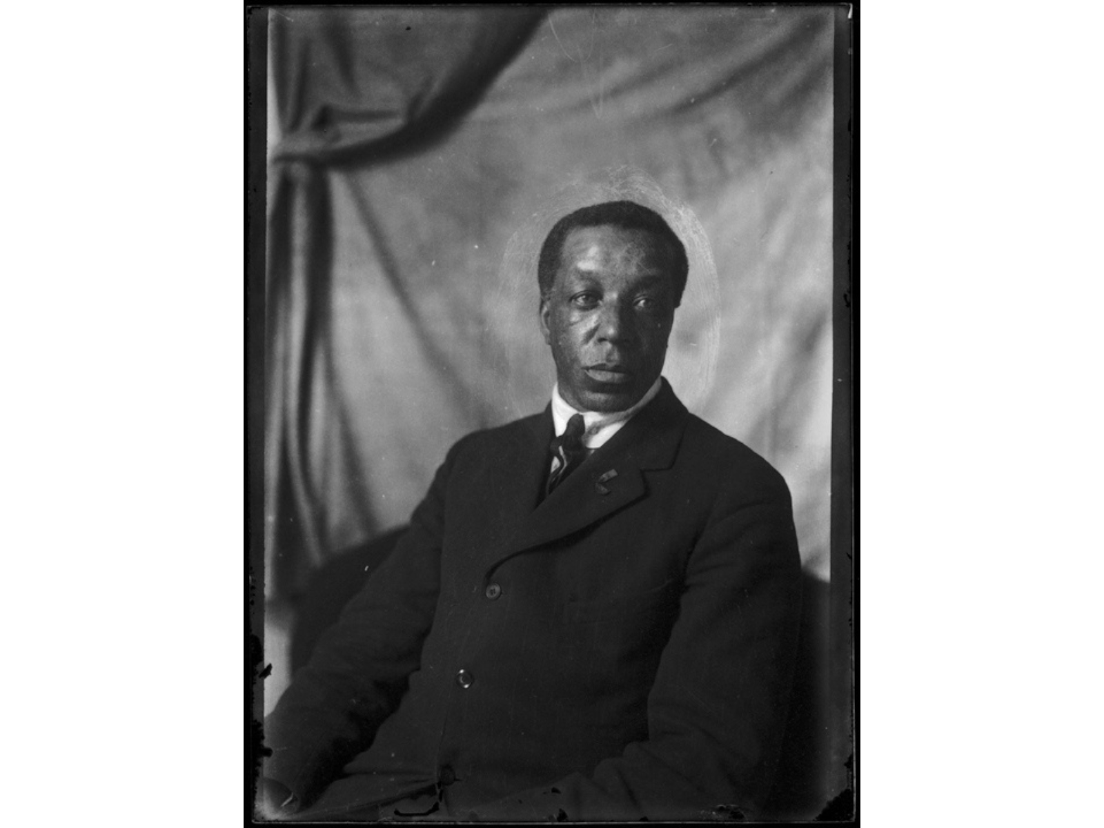 Black and white studio portrait of a man in a dark suit and tie in front of a neutral draping backdrop.