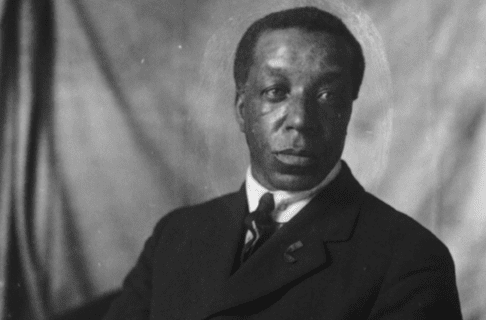 Black and white studio portrait of a man in a dark suit and tie in front of a neutral draping backdrop.