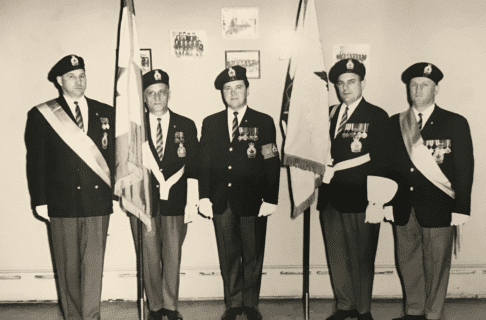Black and white photograph of five men in uniform standing on a row with serious faces. Two of them are holding up flags draped around their poles.