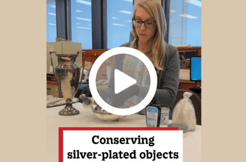 A screenshot of a video, an individual standing behind several silver artifacts in a lab speaks to the camera. There's a play button over the screenshot and overlaid text reads, "Conserving silver-plated objects".