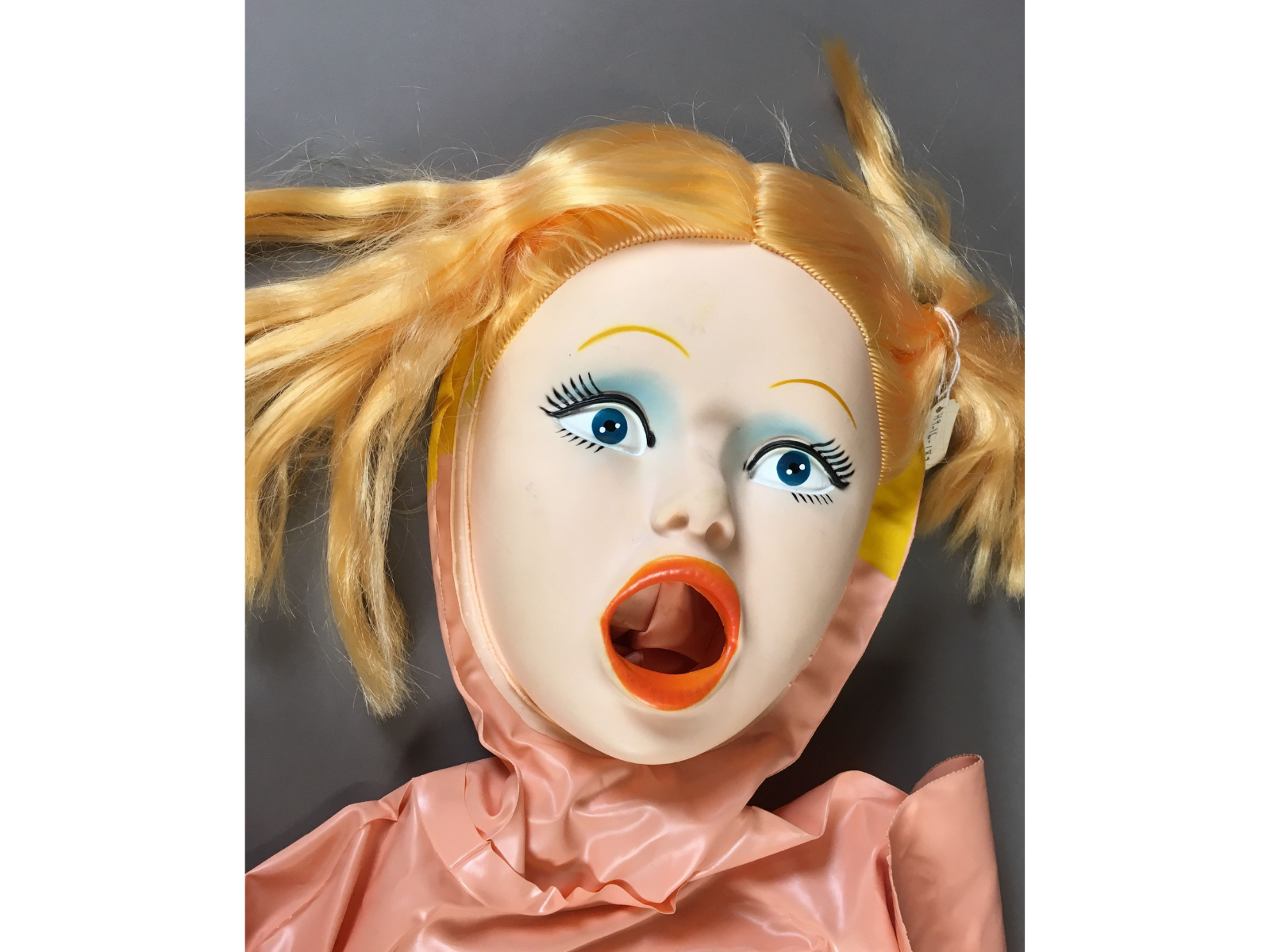 Close-up on the face of an inflatable doll with yellow hair, blue eye shadow, and an open mouth.