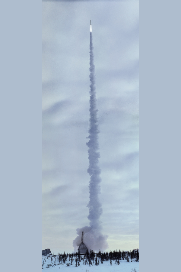 A long plume of smoke rising straight up from the ground to high in a cloudy sky where a rocket launches upwards. On the ground, a pointy building surrounded by evergreen trees on a snowy landscape.