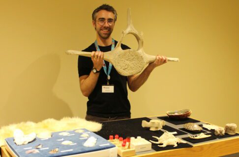 A learning facilitator holds up a whale vertebra above a table of other aquatic specimens.