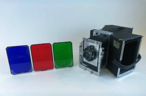 An angular vintage camera with a vaguely triangular-shaped back chamber behind the lens. Beside it blue, red, and green glass plates are displayed.