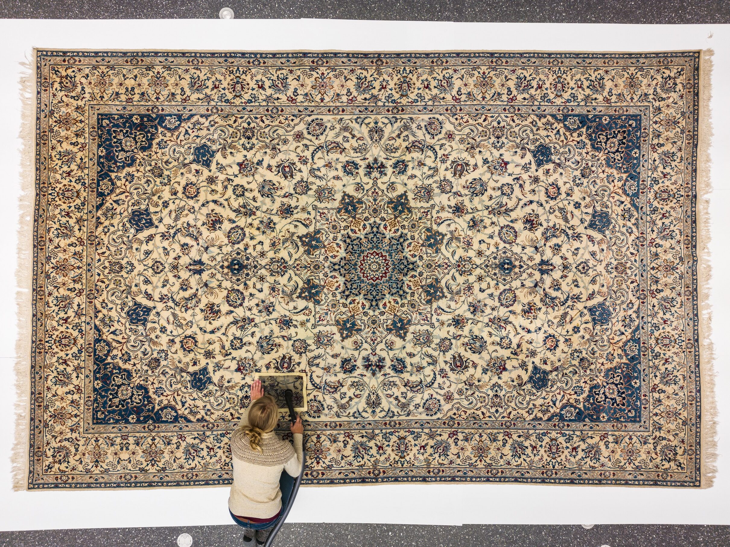 Photograph taken from above. The large Persian carpet has been laid out. Along the lower right edge the Museum conservator kneels, carefully vacuuming a section of carpet through a mesh screen.