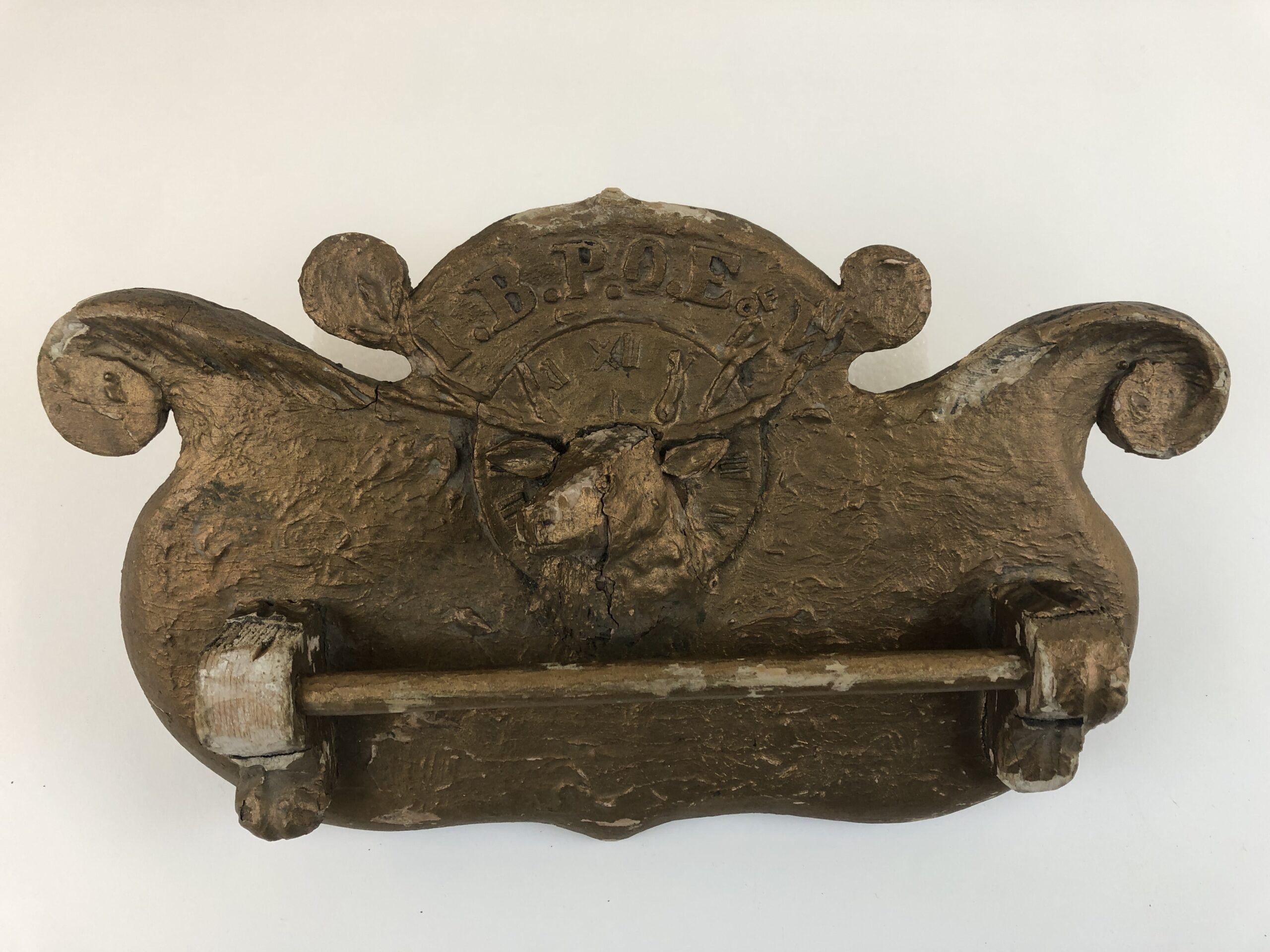 Wood plaque, covered in plaster and painted gold. "I.B.P.O.E. OF W." in raised print across top, an elk head with a clock face in the background.