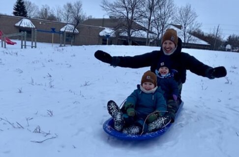 An adult and two children smiling as they ride a sled together down a snowy hill.