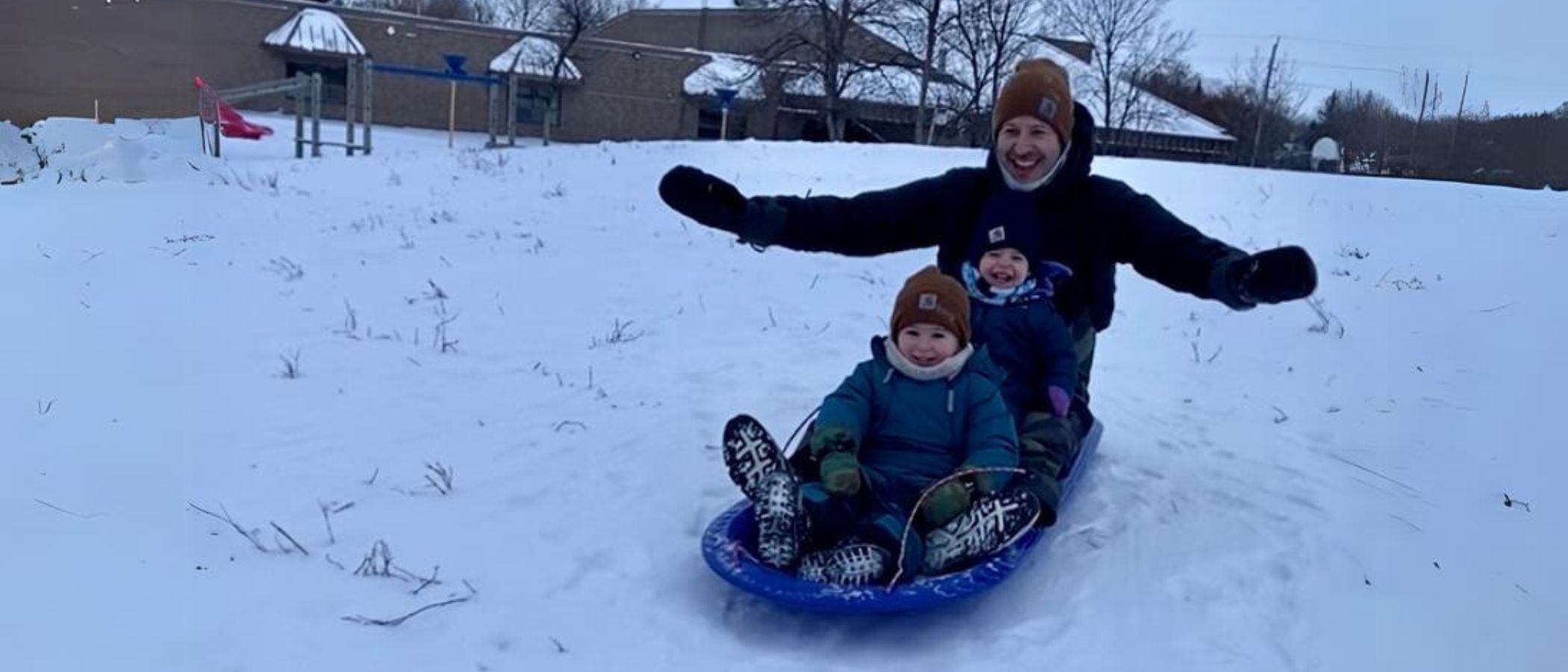 An adult and two children smiling as they ride a sled together down a snowy hill.