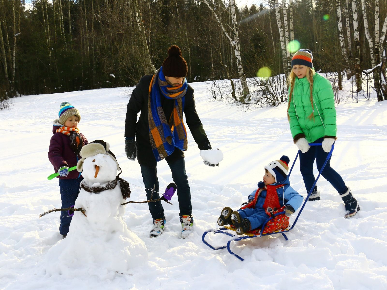 Two adults and two children in winter-wear playing in the snow making snowballs and a snowman.