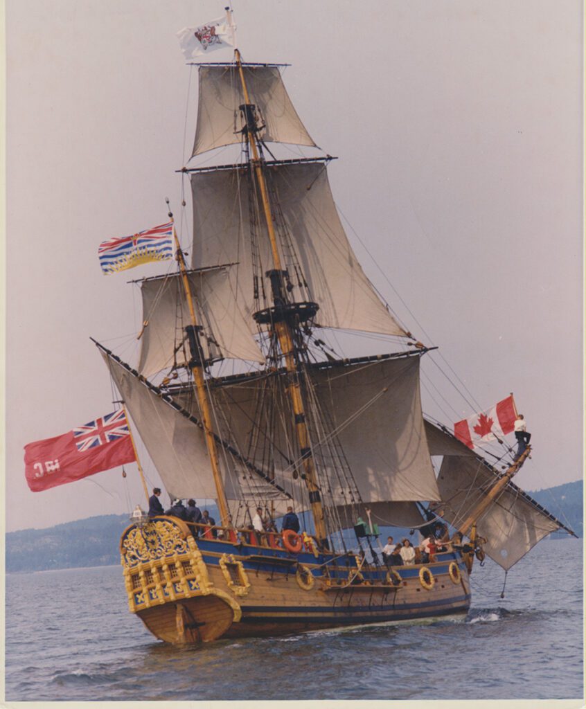 A wooden sailing vessel, the Nonsuch, with full sails and flags raised, sailing on open water. A number of people can be seen standing on the deck.