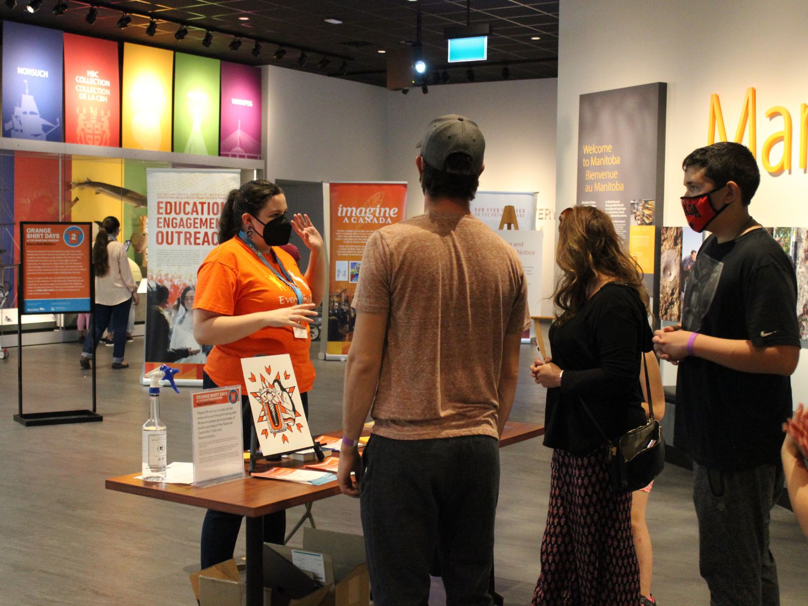 A Museum staff person wearing an orange t-shirt standing behind a table in the Welcome Gallery speaking with three Museum visitors. In the background, further inside the gallery is signage and banners for Orange Shirt Days.