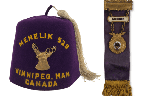 On the left, a purple fez with a white tassel. Embroidered on the fez is an elk’s head and the words, “Menelik 528 / Winnipeg, Man. Canada”. To the right, beside the fez, a purple ribbon fixed to a gold clasp with a gold fringe at the bottom. From the top clasp chain holds up a “Member” badge and a round clock symbol. Faint lettering on the ribbon reads, “Menelik Lodge No 528 / I.B.P.O.E of W. / Winnipeg, Man. Canada”.