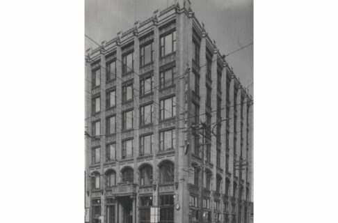 Black and white photograph of a rectangular six-story office building. Accent pillars rise up between each column of windows and there is a grotesque at the top of each, and a grinning head at the bottom.
