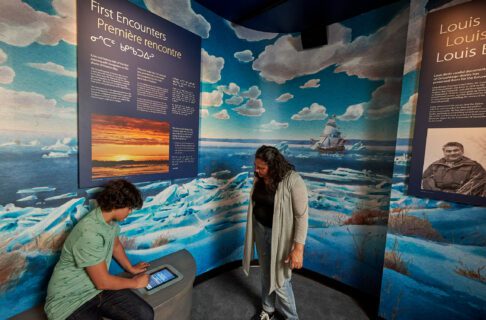 A young visitor is seated on the bench exploring the touchscreen function of the audio experience while a adult looks on. The room is a cozy alcove with a mural depicting ice on Hudson Bay, blue sky and white puffy clouds, and ship off in the distance.