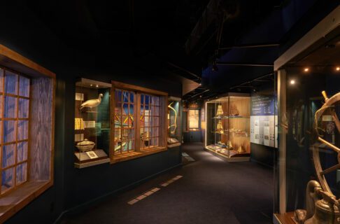 Dark blue walls offset the brightly-lit exhibit cases that feature artifacts related to nautical history and scientific collecting. Wooden framed windows look out over the Nonsuch Gallery.