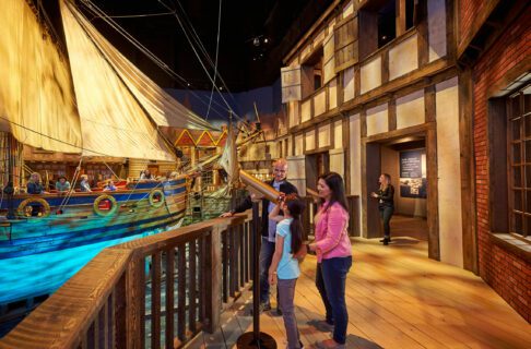 Two adults are standing next to a child who is using a brass telescope to look at the Nonsuch ship.