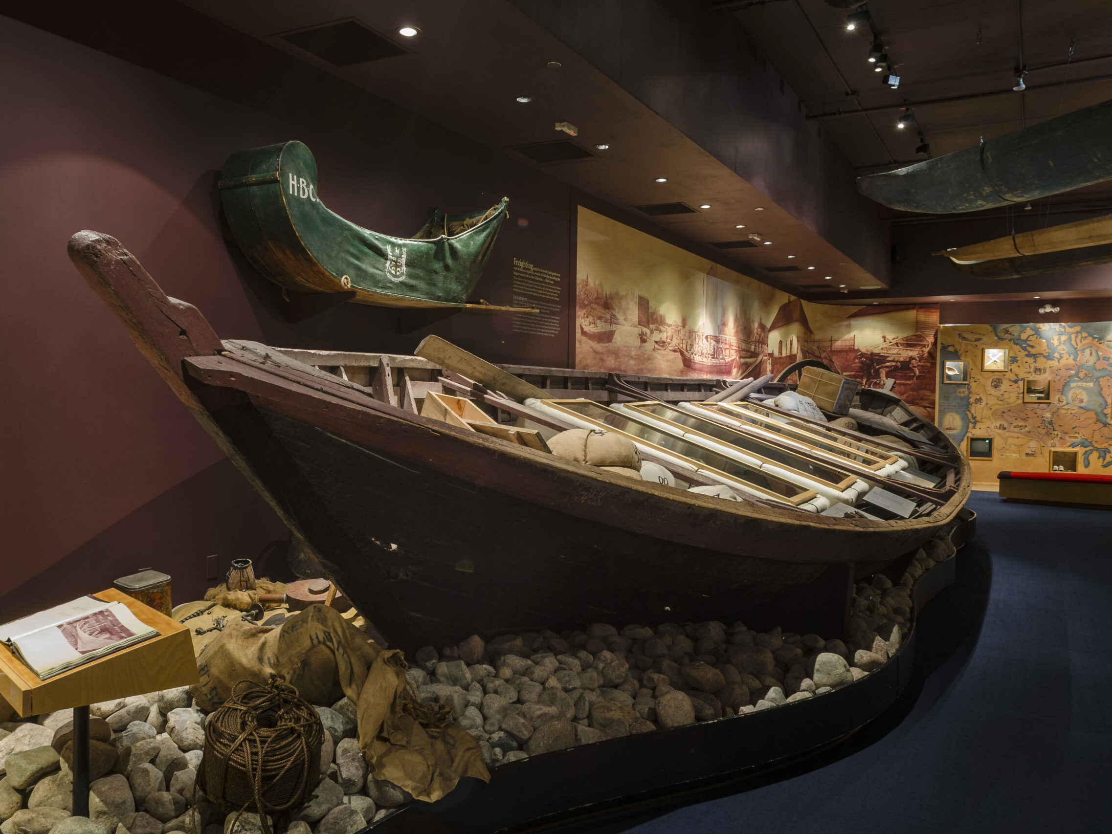 A large York boat is nestled amongst larger rocks with artifact cases inside made to look like fur bales but featuring artifacts related to freighting and transportation. The walls of the gallery are a deep burgundy and there is a mural behind the boat depicting scenes from HBC’s prominent trading post at Norway House.