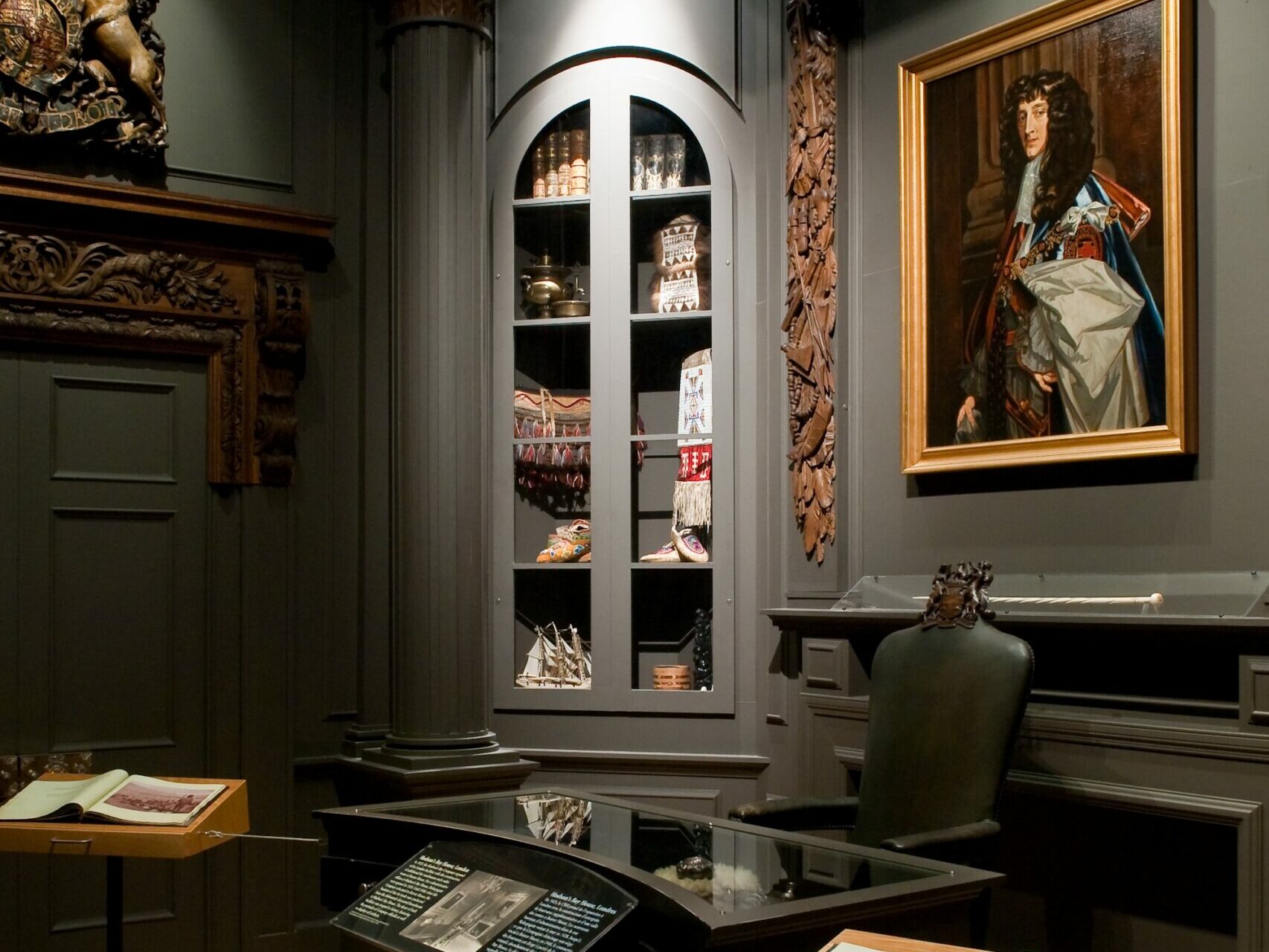 A well-lit area with dark grey walls, fancy columns, and a fake fireplace and mantle. Dark wood carvings from the former HBC head office in London frame a colourful oil painting of Prince Rupert above the mantle. A leather chair sits at an exhibit case made to look like a desk, and two curatorial notebooks with black and white images flank the desk.