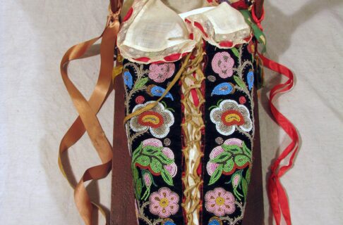 A flat piece of wood supports an oblong-shaped bag made from black velvet and heavily decorated with glass beads in floral patterns. Colours of light pink, bright green, blue, red, yellow, and white are featured in the design. A curved piece of wood runs along the top, called a fender, and gold and red ribbons are tied to the side.