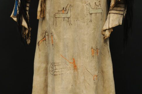 A tan-coloured hide shirt placed on a mannequin that has painted depictions of people with horses, possibly representing battles, using dark brown and bright orange colours. A large quillwork panel in the centre with a geometric pattern and two strips of geometric quillwork along the shoulders. Pieces of dark brown horsehair dangle from the collar and shoulders.