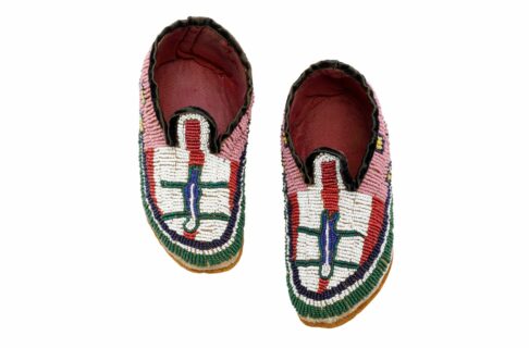 A pair of child’s moccasins with toes pointing down. The insides are lined with pink fabric, and the exterior is covered in brightly coloured glass beads. The beads are pink near the ankle, the vamp has white and red along with a figurative image in the centre, and the edges are lined with rows of blue, white, and green.