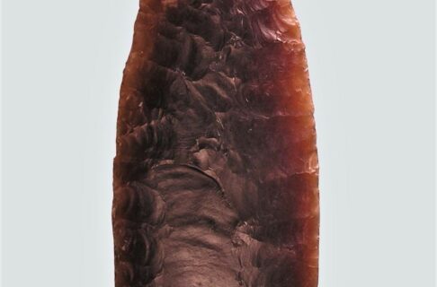 A spear head made from translucent brown flint. Flakes of stone have been removed from its surface, and a long flake removed from its base halfway to its tip.