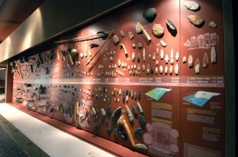 Hundreds of archaeological artifacts attached to a gallery wall, including spears, arrowheads, axes, and ceramic sherds.