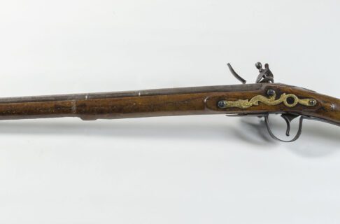 A long rifle with the barrel pointed to the left of the image in order to show the brass serpent side plate above the trigger. Part of the flintlock mechanism can be seen over the top of the rifle.
