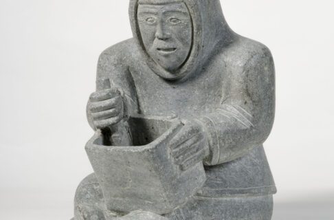 A light grey soapstone carving depicting a seated man wearing a parka with a pointed hood, holding a large rectangular soapstone pot on his lap while he carves out the interior.