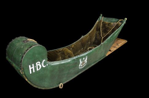 Sled (called a cariole) covered with green painted canvas and interior lined with plush olive-green velvet. The letters HBC are painted in white on the curved front, and the Company’s Coat-of-Arms painted on the side in white and red. The Coat-of-Arms features two elk flanking a crest with St. George’s cross in red and four little beavers in each quadrant, topped with a fox sitting on a cap, and a banner with their Latin motto Pro Pelle Cutem scrolled along the bottom.