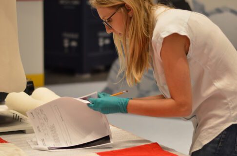 Woman wearing a blue shirt and glasses; holding a notepad and pencil assessing a hockey jersey.