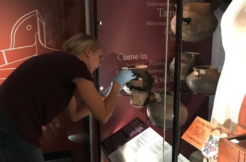Woman wearing a purple shirt and blue gloves, placing an archaeological pot into an exhibit case.