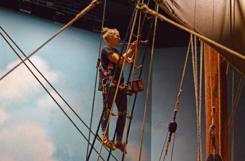 Woman wearing a pink harness and holding a paint brush with tar, on the Nonsuch rigging.