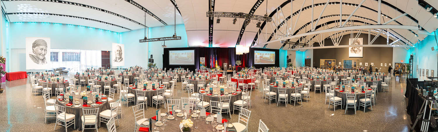 Panoramic view of Alloway Hall, a large event hall with high, white ceilings, decorated for an event with laid tables and a stage set up.