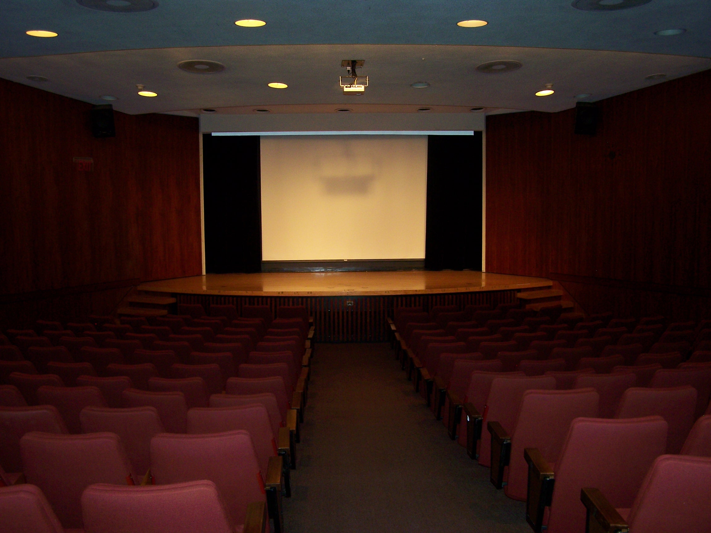 View down the central aisle of a theatre style auditorium. Either side are rows of red, plush chairs. At the front is a raised wooden stage, and a projector screen hung against a black background.