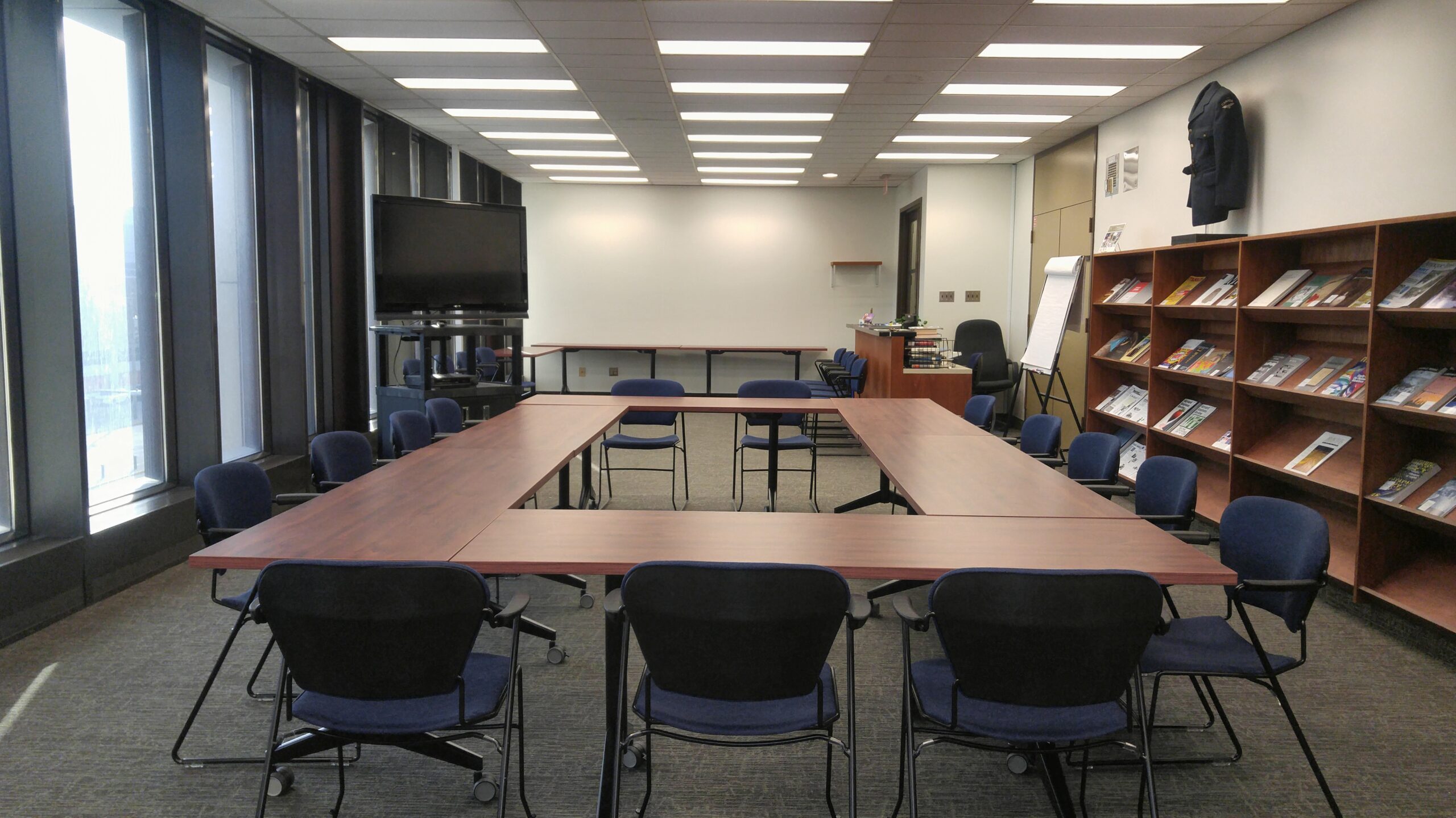 An office boardroom with tables lined up together in a rectangle in the centre of the room. On the left side are floor-to-ceiling windows and a large flat-screen television on a rolling cart. To the right are book rack-style shelves.