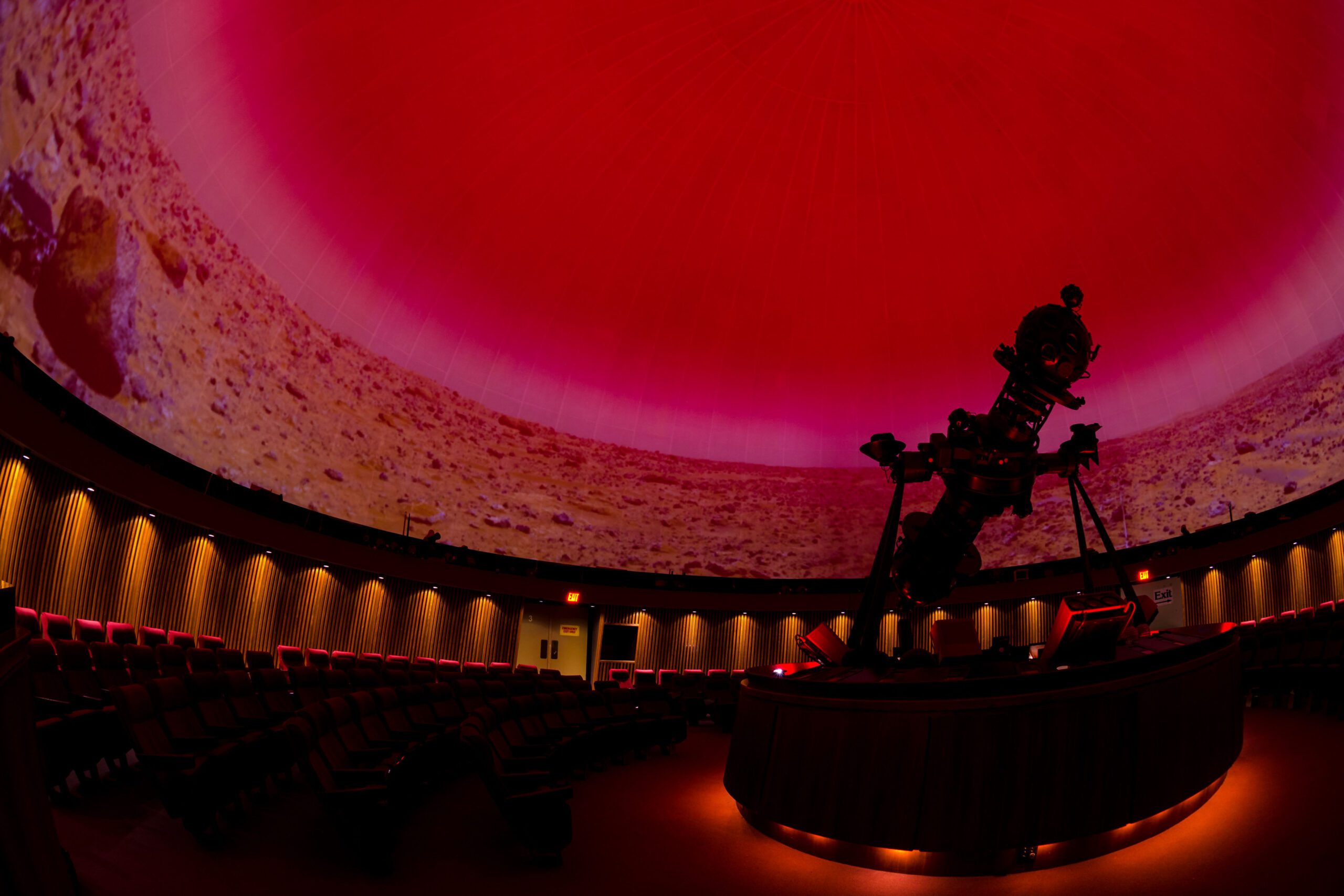 A wide-angle view of the inside of the Planetarium. On the right side is the Zeiss star projector on its platform, and displayed on the domed screen is a dry, rocky landscape and a red sky.