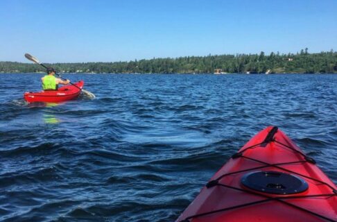 Photo taken from in a red kayak on a lake. Ahead, to the left a kayaker in another red kayak, wearing a bright green life jacket, paddles forward. In the background, the shore is wooded.
