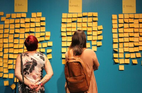 During Orange Shirt Days at the Manitoba Museum, two individuals reading orange sticky notes arranged on a teal wall under headings reading, "I feel / I learned / I will".