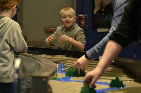 A smiling child engaging with the Science Gallery water table as someone from out of frame places a plastic tree in the path of the water.