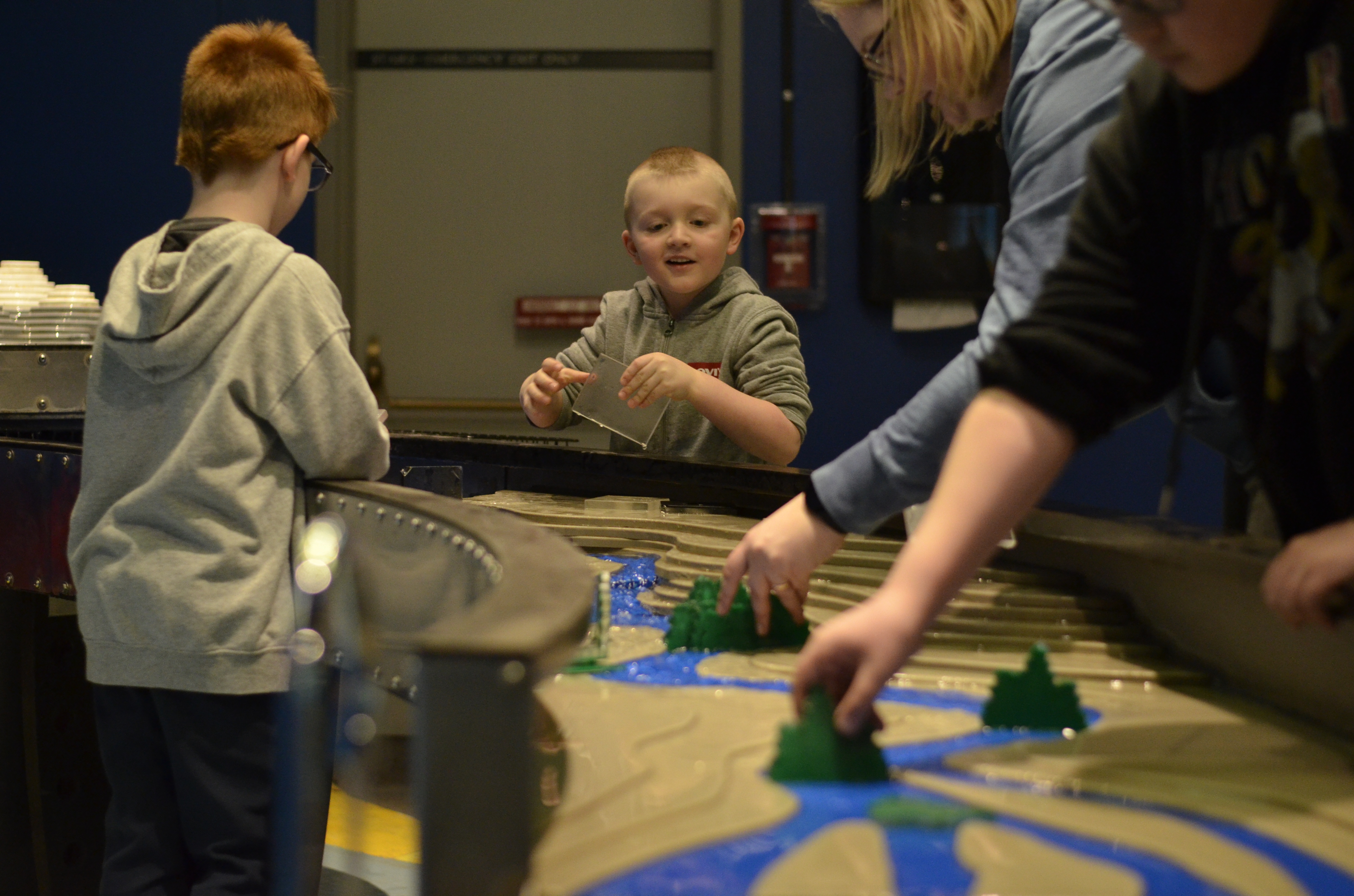 A smiling child engaging with the Science Gallery water table as someone from out of frame places a plastic tree in the path of the water.