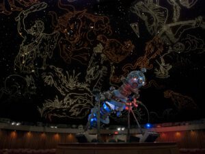 Interior of the Manitoba Museum Planetarium. A large Zeiss star projector on a central platform lit in blue and red. The domed screen behind is illuminated with illustrations outlining the constellations in the stars.
