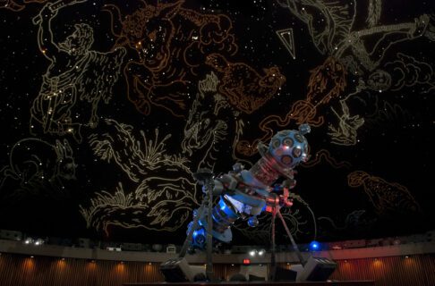 Interior of the Manitoba Museum Planetarium. A large Zeiss star projector on a central platform lit in blue and red. The domed screen behind is illuminated with illustrations outlining the constellations in the stars.