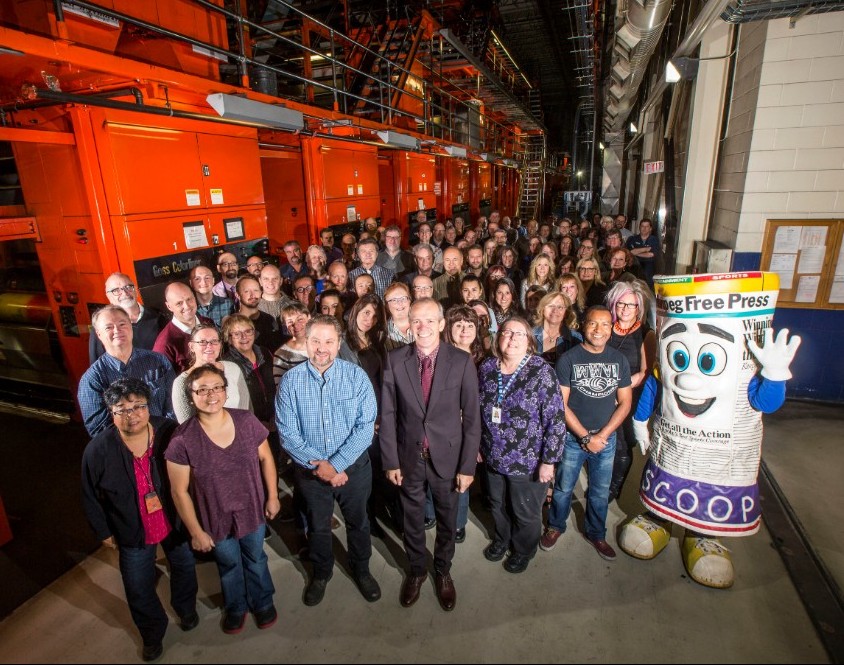 A large group of the Winnipeg Free Press staff with WFP mascot smiling for a group photo in the printing room.