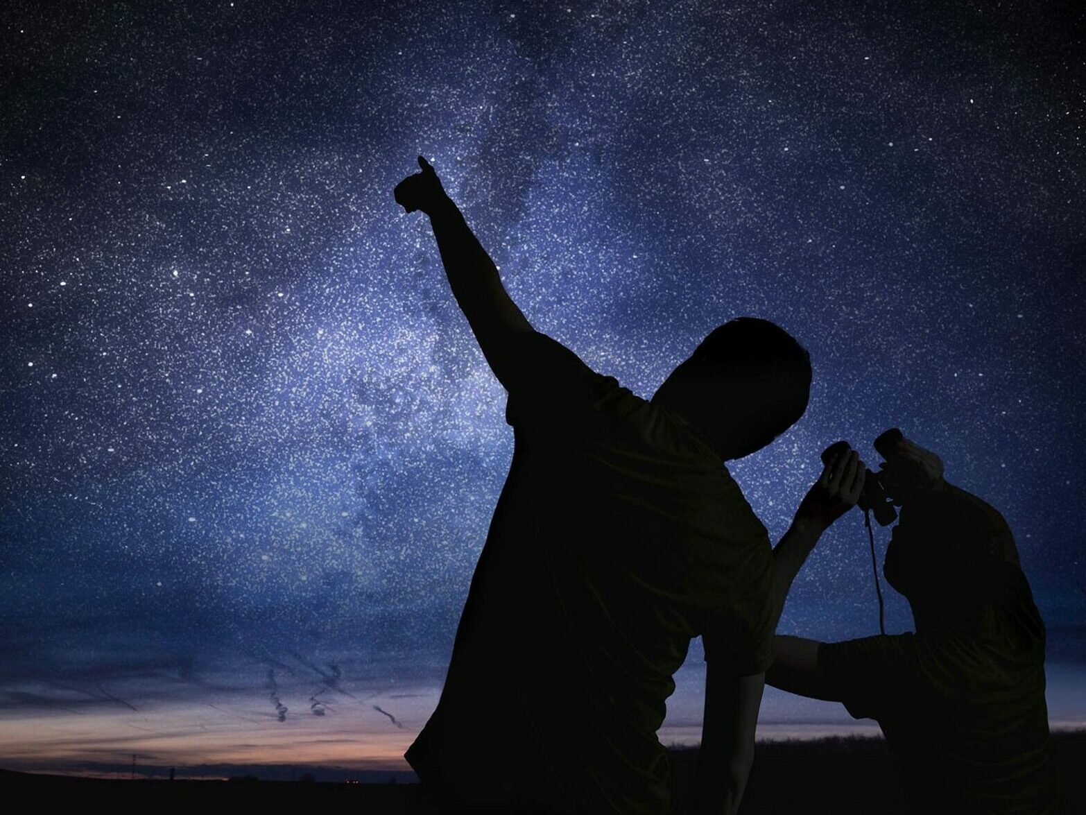 Silhouettes of an adult and child. The adult points up into the night sky full of stars, as the child looks up through binoculars.