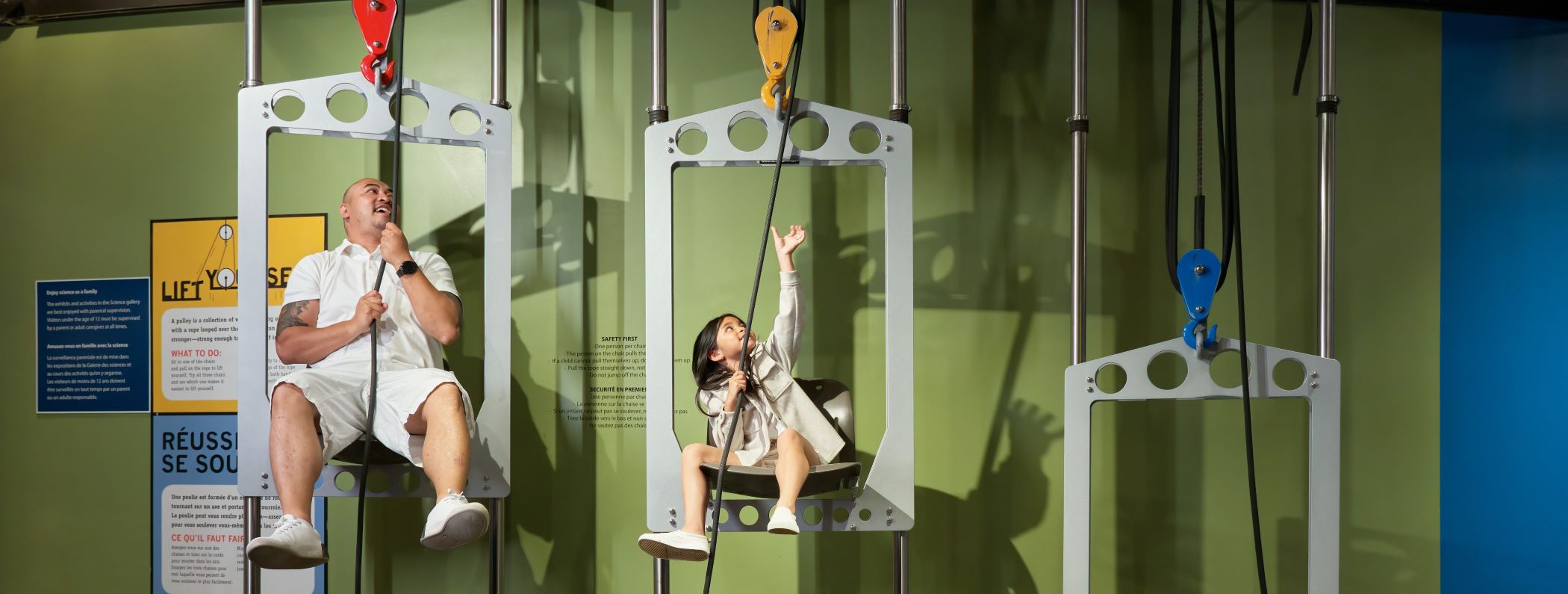 An adult and child race on the Pulley Chairs in the Manitoba Museum Science Gallery.